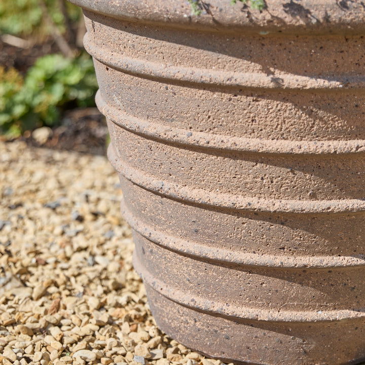 A close-up of a ribbed terracotta-colored plastic or concrete planter pot sitting on gravel outdoors.