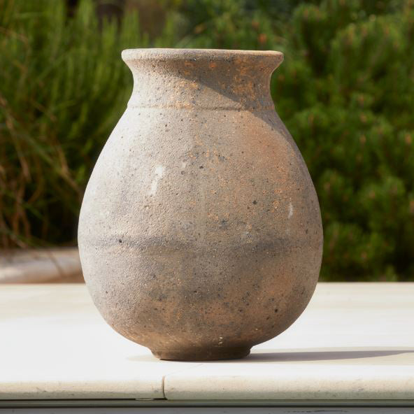 A weathered, rustic stone plant pot with a rounded belly and narrow neck, displayed outdoors on a light surface with green foliage blurred in the background.