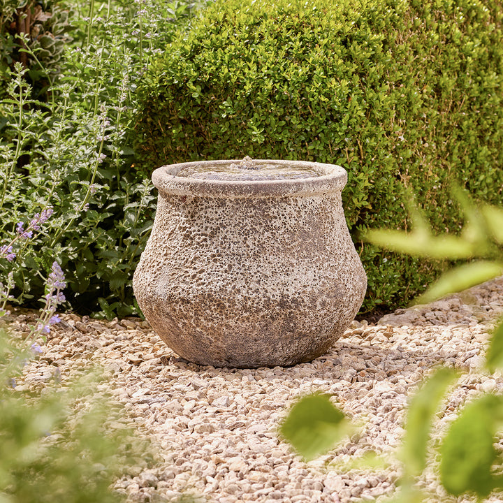 Decorative stone water feature on a gravel surface with greenery in the background