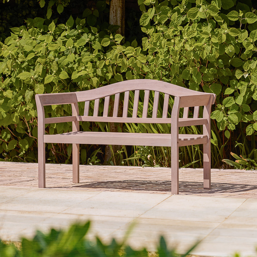 Natural teak effect wooden garden bench with traditional curved backrest and arms, styled on a small garden patio.
