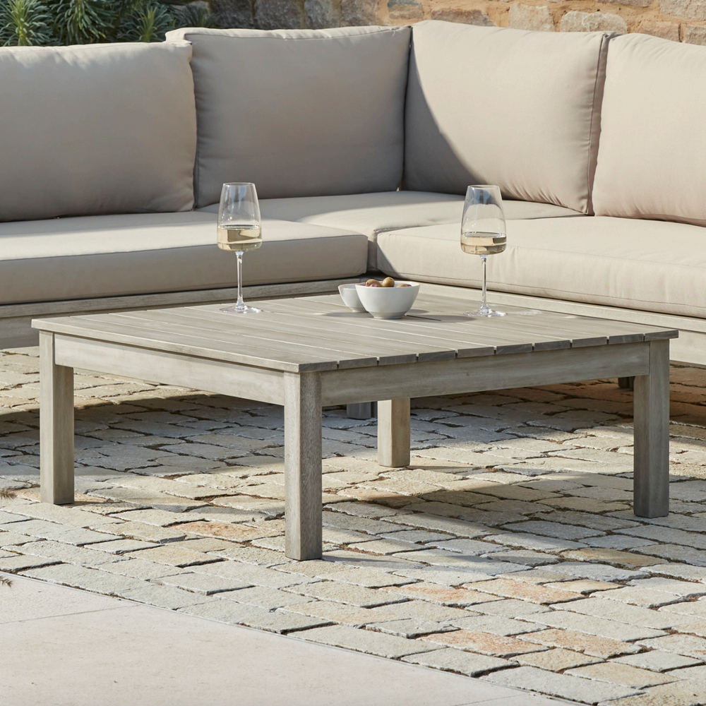 Repton Classic Wooden Garden Coffee Table with a slatted design, part of the Repton Corner Garden Furniture Set. Styled on a stone patio with two glasses of white wine and a bowl of snacks, this weather-resistant outdoor table complements modern patio and garden seating areas.