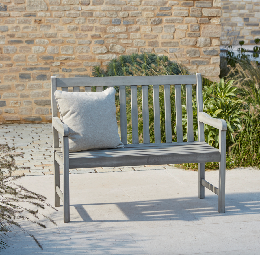 Repton Wooden Garden Bench with a textured grey finish, placed by a rustic stone wall surrounded by blooming greenery. A classic farmhouse-style two-seater bench designed for patios and gardens.