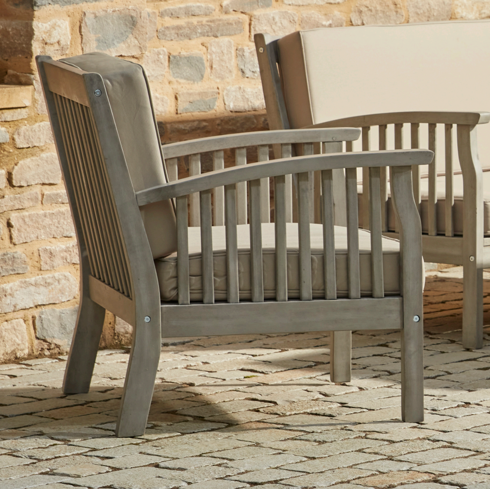 Close-up of the Repton Wooden Lounge Armchair, featuring a slatted backrest, curved armrests, and a weathered grey finish. The chair includes a plush beige cushion for added comfort, making it an elegant and durable choice for outdoor patio and garden seating. Positioned against a rustic brick wall on a stone-paved surface.
