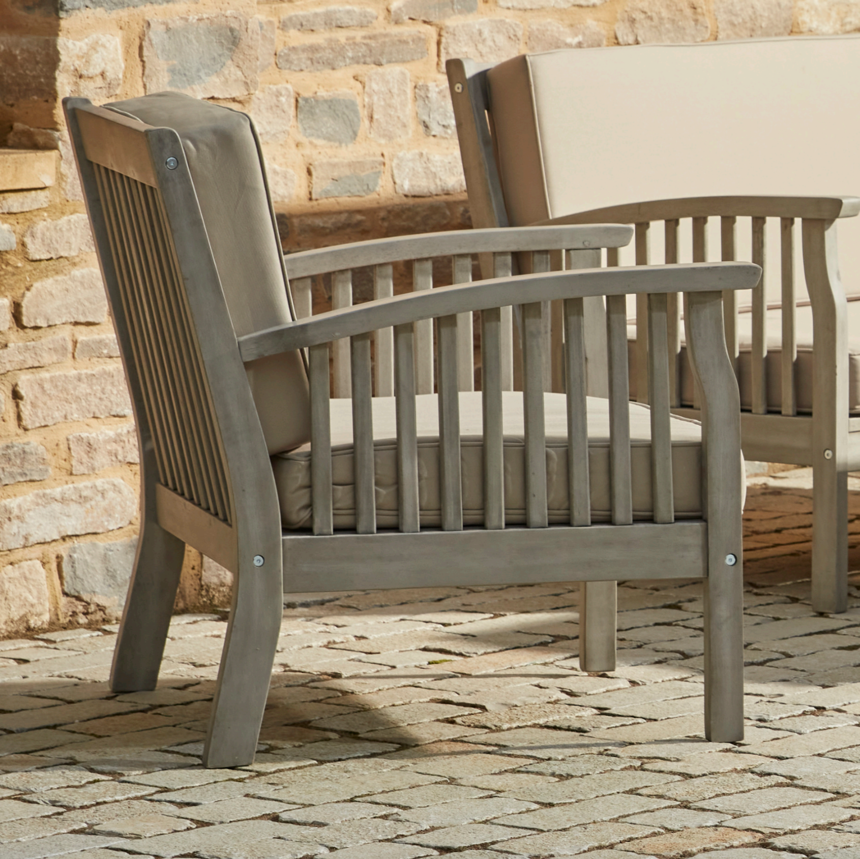 Close-up of the Repton Wooden Lounge Armchair, featuring a slatted backrest, curved armrests, and a weathered grey finish. The chair includes a plush beige cushion for added comfort, making it an elegant and durable choice for outdoor patio and garden seating. Positioned against a rustic brick wall on a stone-paved surface.
