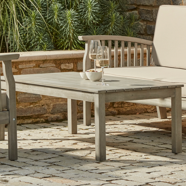 Close-up of the Repton Wooden Coffee Table, part of the Repton Lounge Set, featuring a slatted tabletop and a weathered grey finish. Styled on a stone patio with two wine glasses and snack bowls, this sturdy outdoor table is designed for garden lounging and patio relaxation. Complemented by matching wooden seating with beige cushions.