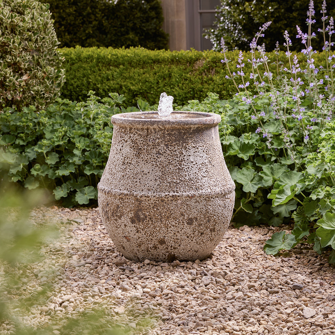 Decorative tall stone water fountain in a garden setting with plants and gravel.