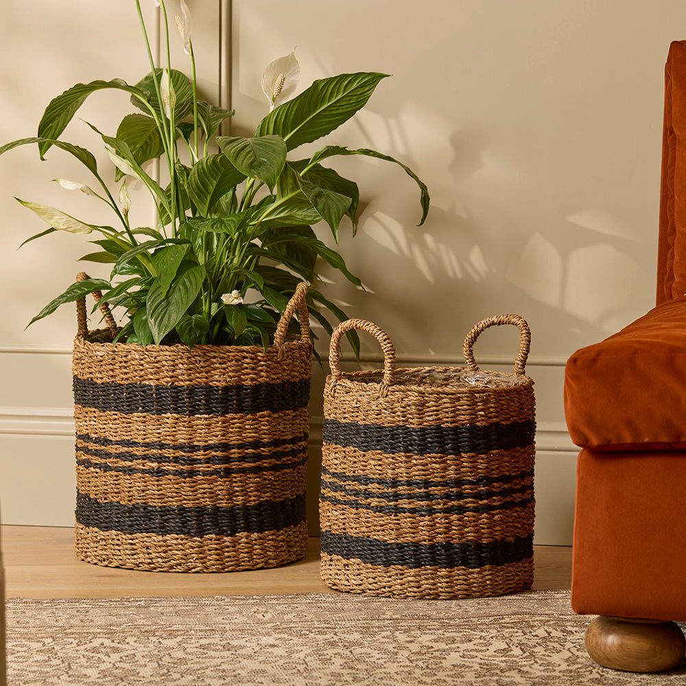 Two woven baskets with striped patterns on a wooden floor next to a plant and a brown chair.