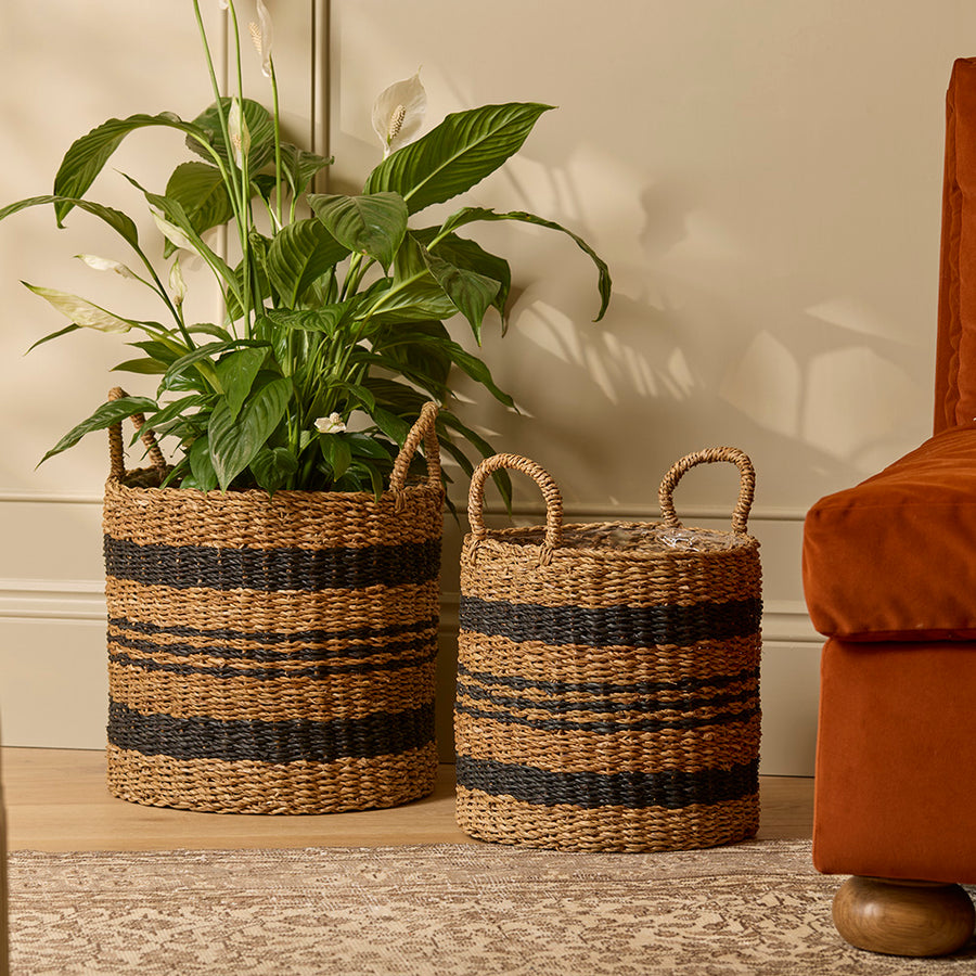 Two woven baskets with striped patterns on a wooden floor next to a plant and a brown chair.
