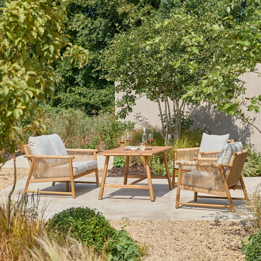 A 4-piece outdoor patio dining set featuring two woven armchairs with light gray cushions, a wooden casual dining table, and a matching wooden sofa, arranged on a stone patio in a landscaped garden.