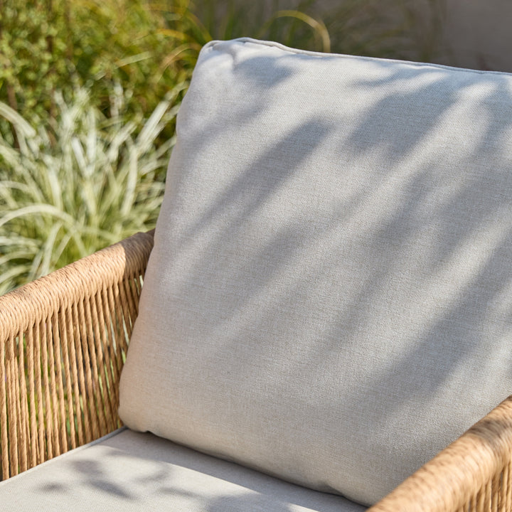 A light beige outdoor patio cushion resting on a woven wicker chair, with dappled sunlight and green garden foliage softly blurred in the background.