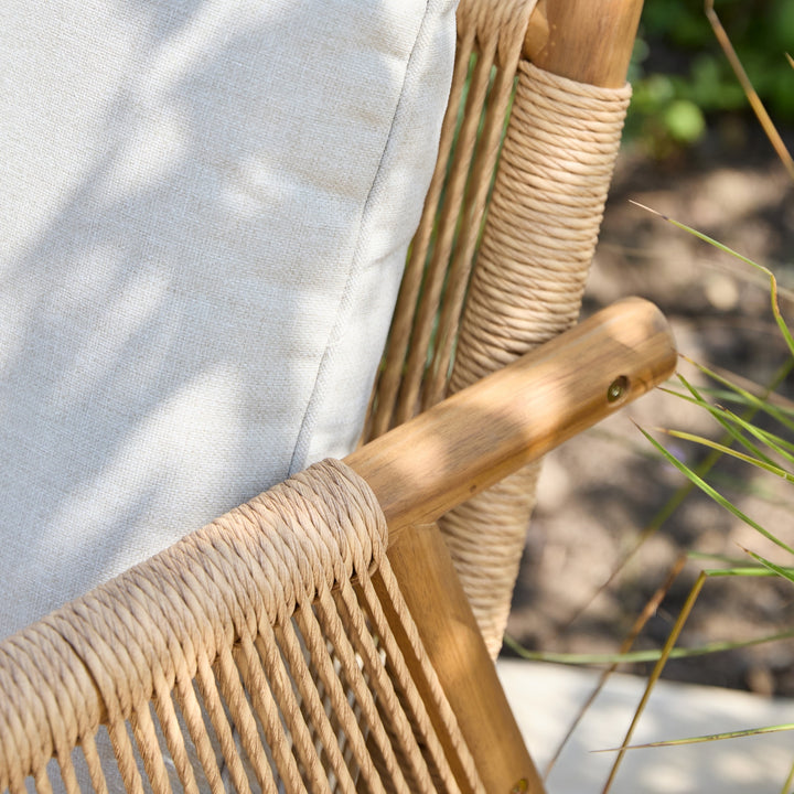 Close-up of a woven outdoor chair with a light beige cushion, natural wood frame, and rope-wrapped armrests in a sunlit garden setting.