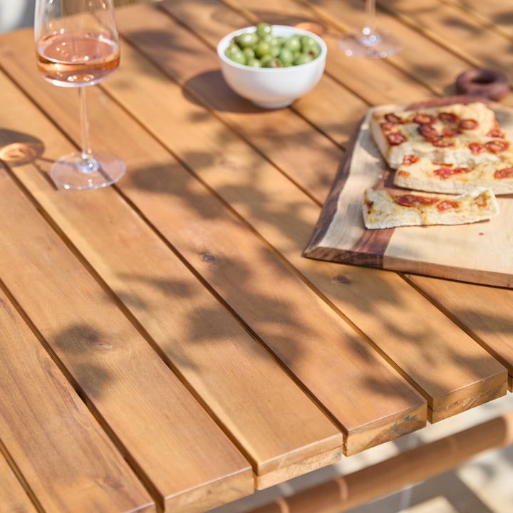 A warm-toned wooden outdoor dining table with parallel slats, dressed with wine glasses, a bowl of green grapes, and pizza on a cutting board under dappled sunlight.