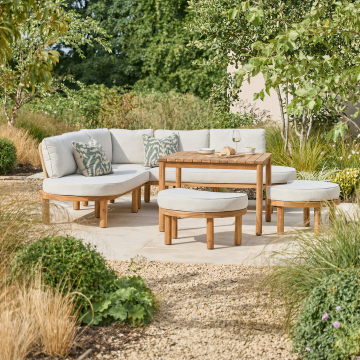 Outdoor patio corner sofa set with a curved wooden frame, rectangular coffee table, and two rounded ottomans with light beige cushions, arranged on a stone patio in a landscaped garden.