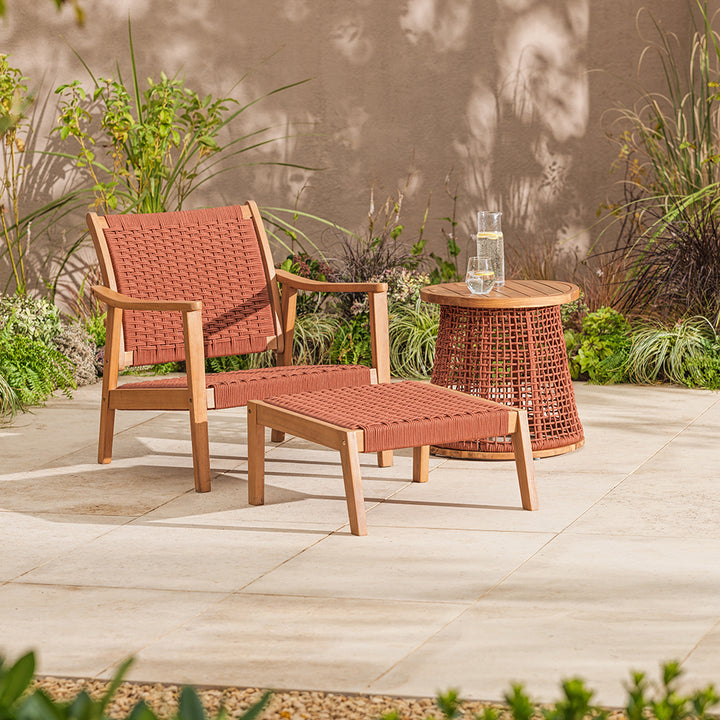 Outdoor patio set with wicker chair, ottoman, and table on a stone patio with plants in the background.