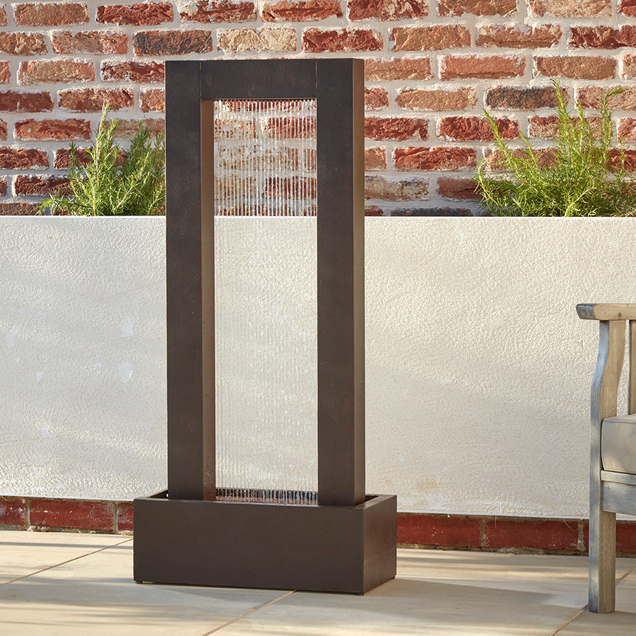 A tall, steel waterfall sat on a buff coloured patio. The water is dropping like raindrops, illuminated by a warm led light