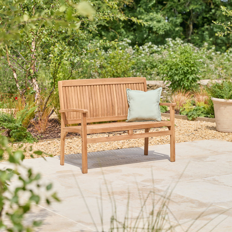 A wooden outdoor bench with armrests sits on a stone patio in a garden setting, with a light sage green throw pillow resting on the seat.