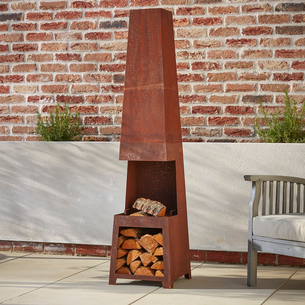 A tall rustic steel chimenea and log store, filled with kiln dried logs ready to burn. Nestled in a corner of the patio next to a wooden garden chair