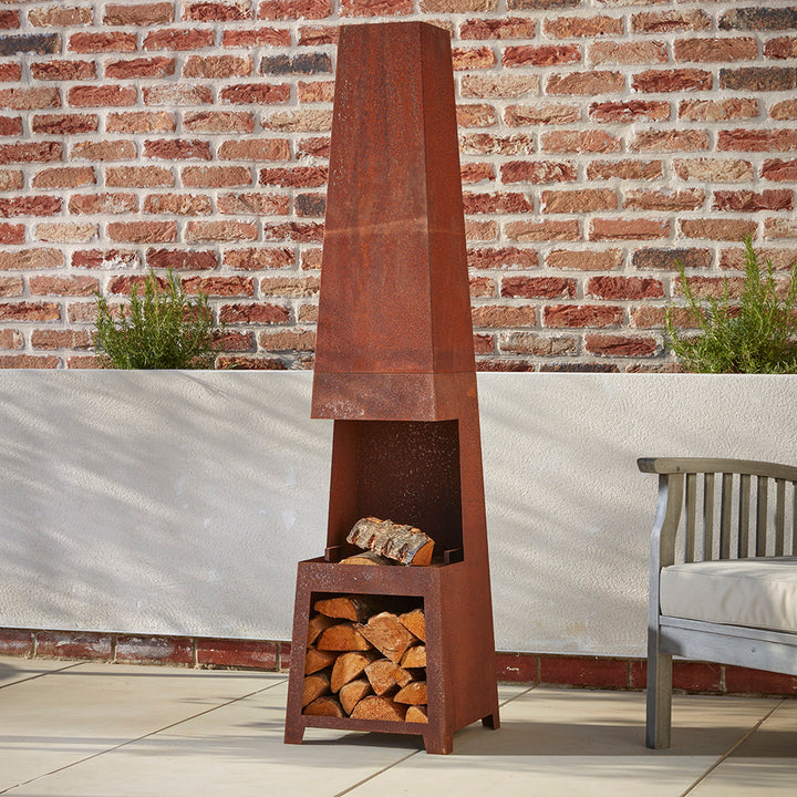 A tall rustic steel chimenea and log store, filled with kiln dried logs ready to burn. Nestled in a corner of the patio next to a wooden garden chair