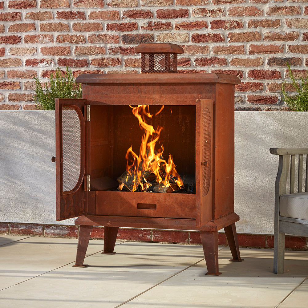 Wakehurst Outdoor Wood Burning Fireplace with its mesh doors open, displaying a strong burning fire. A durable steel garden heater with a rusted finish for a vintage aesthetic.