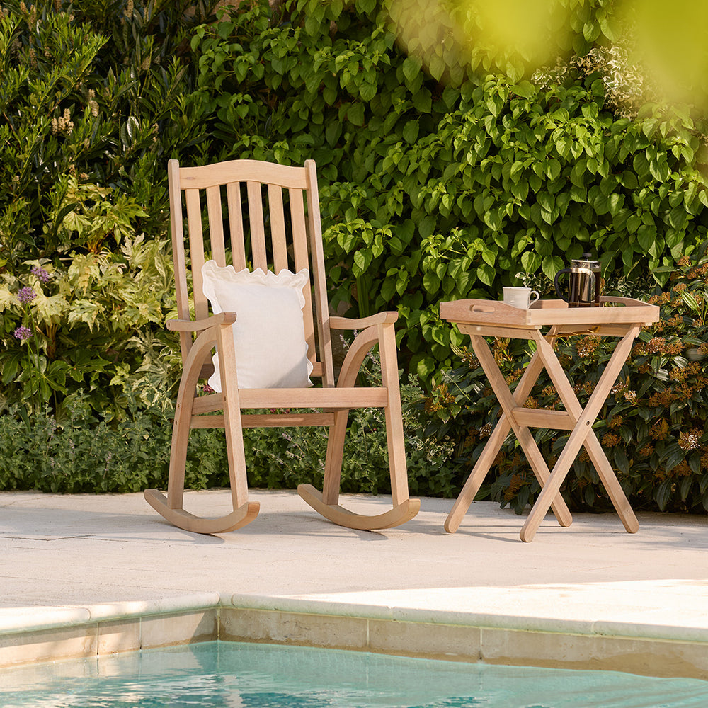 Wooden rocking chair and side table by a pool with greenery in the background