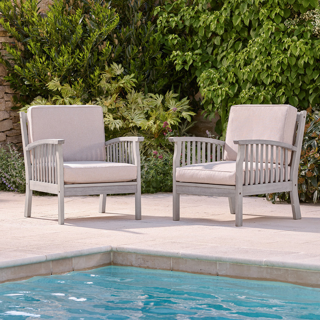 Pair of grey wooden outdoor armchairs with oatmeal cushions, placed by a pool with green garden backdrop.