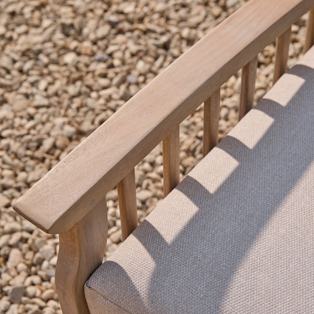 Detail of smooth wooden armrest on natural outdoor armchair with oatmeal cushion, set against gravel patio.