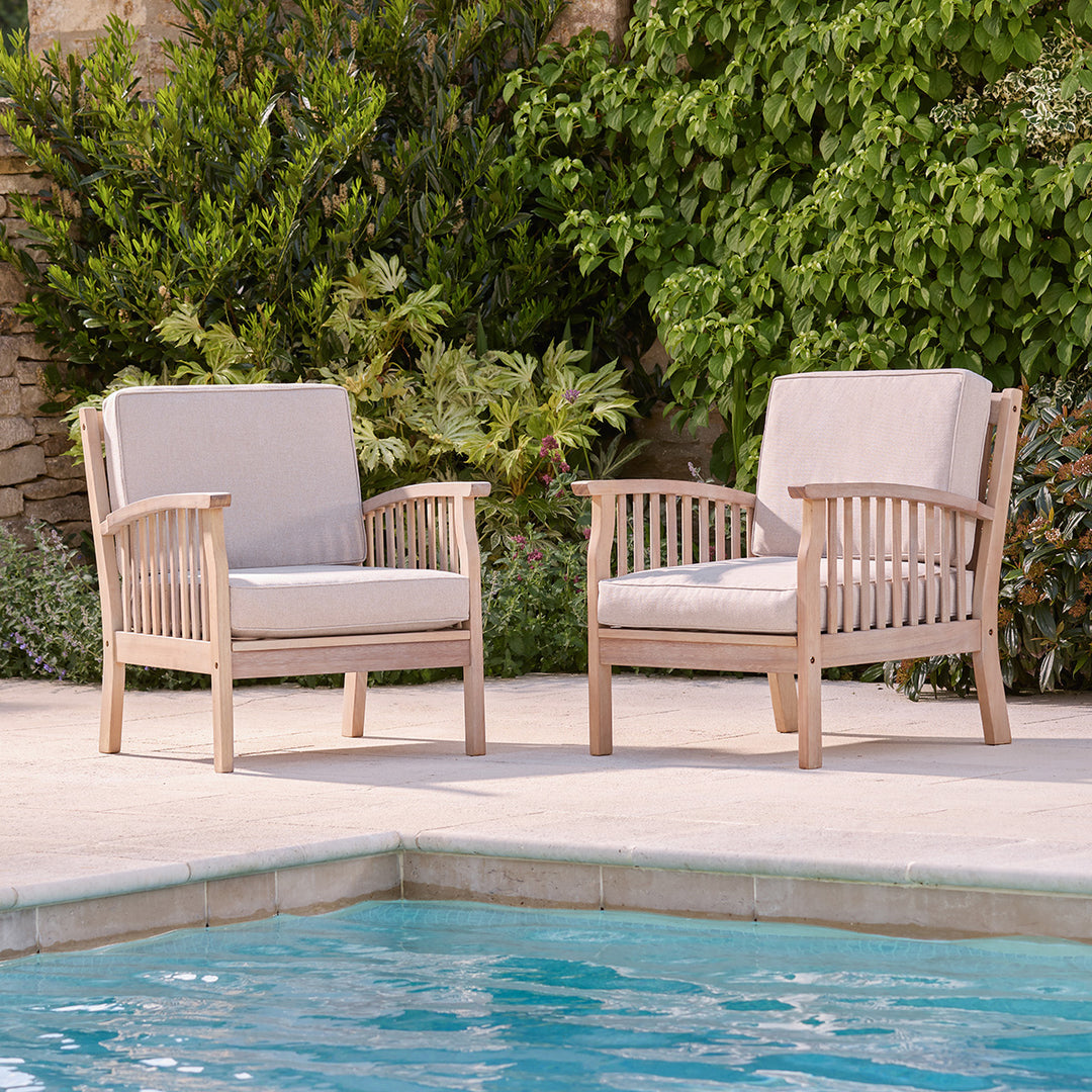 Pair of natural wooden outdoor armchairs with oatmeal cushions, styled poolside with garden backdrop.