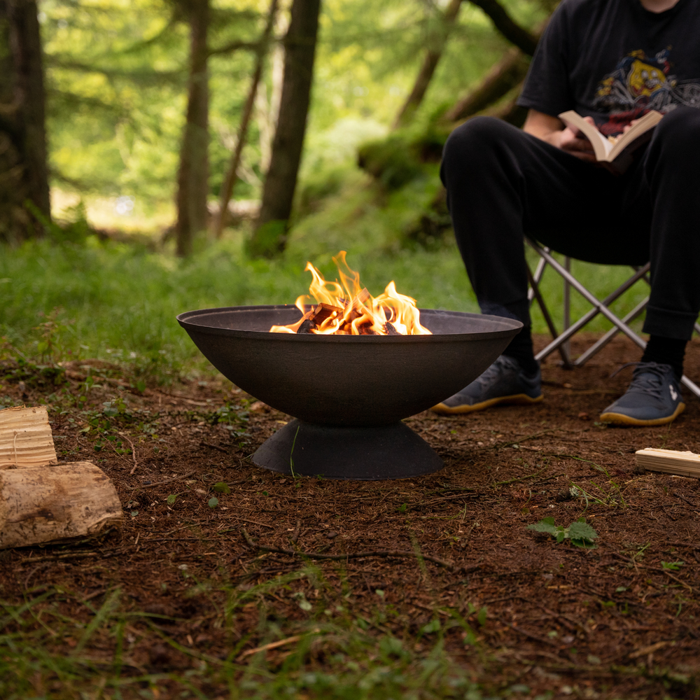 A fire pit burning in a woodland setting with a person sitting in a camping chair nearby, enjoying the warmth.